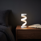 White spiral LED table lamp lit on a walnut bedside table beside an open book
